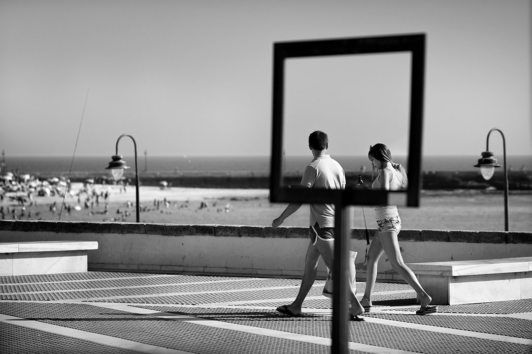 Two people walk behind a rectangular frame beside the beach.